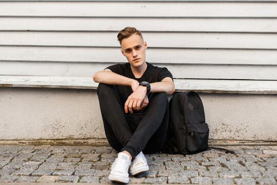 Handsome fashion man model in fashionable black clothes with a T-shirt and pants with white sneakers with a backpack sits near a wooden building