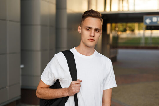 young handsome man model in a fashionable white T-shirt with a backpack walks on the street and straightens his hair