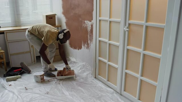 Side full footage of African American man painting walls with brown color using paint roller and listening to music in headphones in new house