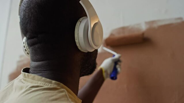 Rear closeup of African American painter in white headphones listening to music while applying lacquer by paint roller in new apartment