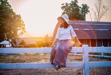 A serene young woman in a sun hat sits on a white fence, soaking in the warm glow of a countryside