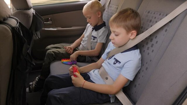 Two little boys are sitting in the backseat of a car wearing seat belts and playing with an anti-stress toy pop it. 