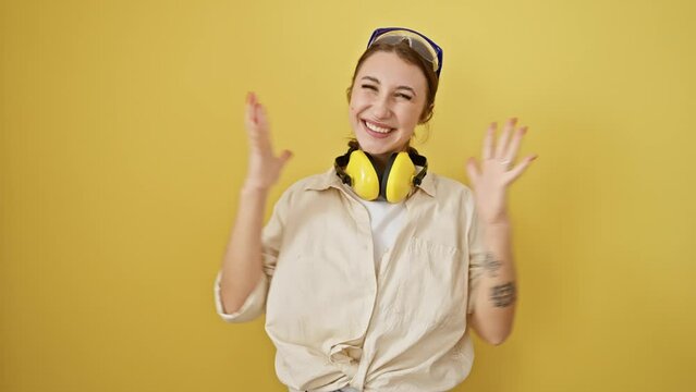 Young brunette girl wearing safety glasses and noise reduction headphones celebrating victory with happy smile and winner expression with raised hands over isolated yellow background