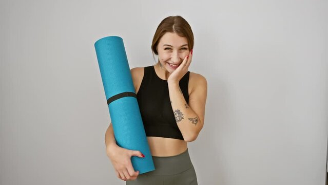 Young brunette girl in sportwear, joyfully celebrating her yoga win with a confident smile and triumphant expression, holding mat against a white isolated background.