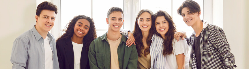 Banner with a group portrait of happy diverse teenage friends. Smiling multiracial boys and girls in casual clothes standing together by a white window. Friendship concept