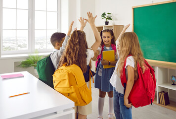 Small group of happy cheerful school children with colorful backpacks standing in a modern classroom and giving each other high five. Back to school, education, success, teamwork concepts