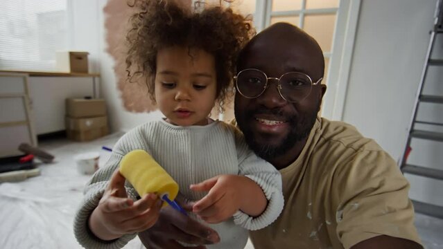 POV UGC footage of African American man in glasses recording blogger content for vlog with his little Biracial daughter who holding mini paint roller and looking at camera during room renovation