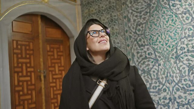 A smiling woman in a black scarf stands in front of an ornate topkapi palace tile wall in istanbul, turkey.