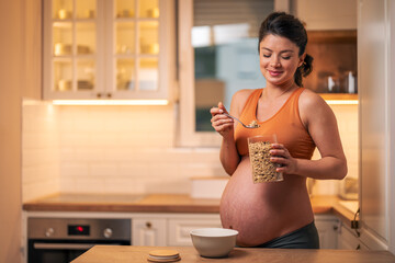 A contented adult pregnant woman in sports outfit having oatmeal for breakfast in the kitchen
