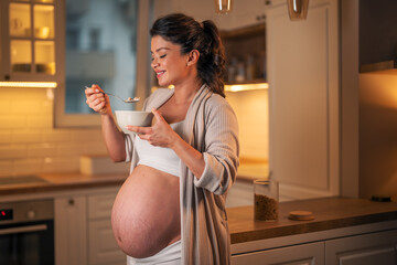 A smiling adult pregnant woman eating a bowl of tasty oatmeal for breakfast in a kitchen