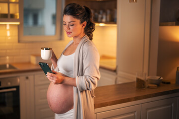 A beautiful mid adult pregnant woman typing on her phone and having a cup of coffee in the morning