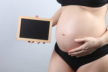 Pregnant woman in underwear holding blank chalk board on gray background. Copy space for information