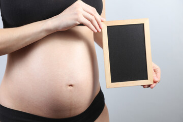 Pregnant woman in underwear holding blank chalk board on gray background. Copy space for information