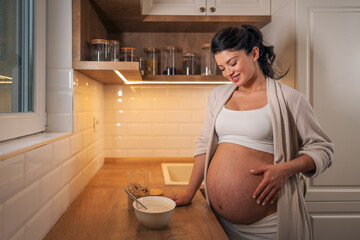 A satisfied pregnant woman looking at and caressing her stomach while enjoying her breakfast in the...