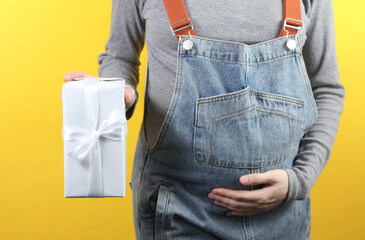 Pregnant woman in denim overalls holding gift box on yellow background