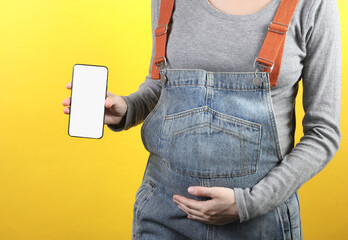 Pregnant woman in denim overalls holding smartphone with white blank screen on yellow background. Space for your information