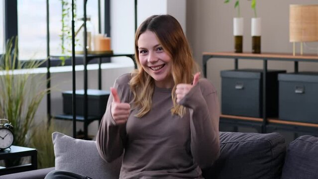 Satisfied Caucasian woman showing thumbs up gesture in agreement with both hands sitting on sofa at home living room. Satisfaction and recommendation.
