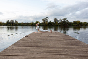Fototapeta premium A mature woman with a short haircut in a white suit is doing yoga on the riverbank. Bhujangasana, Upward Dog