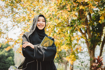 A woman wearing a black abaya and holding a Quran faith The Holy Al Quran book. She is smiling happy outdoor nature, arabic calligraphy translate meaning of Al Quran