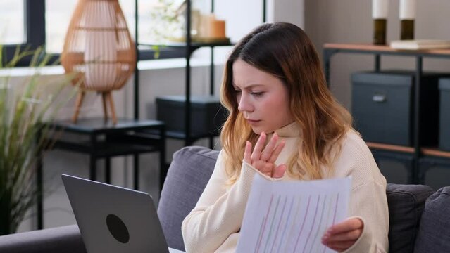 Thoughtful and serious Caucasian female freelancer sitting on sofa, working at home office with documents and laptop. Student studying online.