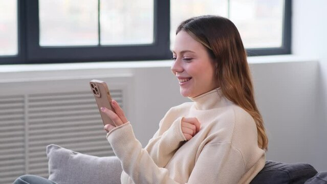 Happy Caucasian young woman laughing while using smartphone at home. Messaging in social media, browsing internet. Spending leisure time with online entertainment.
