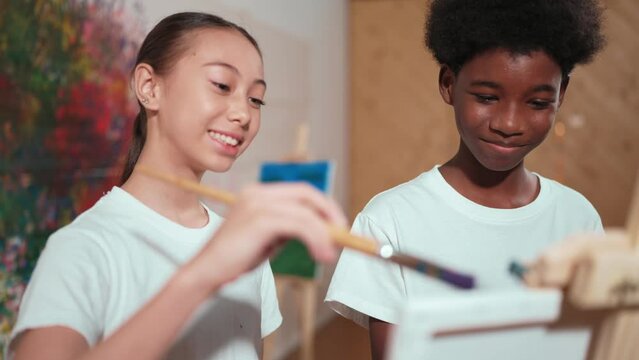 Diverse children in casual shirt painting canvas together in creative art lesson. Multicultural happy high school student drawing a same canvas while standing at colorful stained wall. Edification