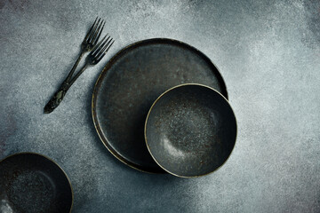 An aesthetic arrangement of vintage dark bowls, plates and cutlery on a gray concrete background. Free space for text.