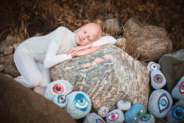 Young hairless girl with alopecia in white futuristic costume sleeping sweetly in surreal landscape with many eyes stones, blurring lines between reality and imagination