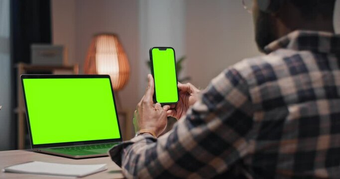 African American man working at home. Man holding smartphone with green chroma key screen. Male using two devices with green template screen. Man looking at green chroma key laptop.