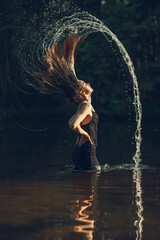Sexy woman in dress throwing wet hair while standing in water of lake in forest at summer sunset  