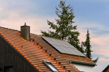 Photovoltaikanlage auf rotem Hausdach vor Bäumen und Abendhimmel - obrazy, fototapety, plakaty