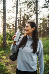 Young caucasian woman pouring and drinking water in forest while taking a break from hiking	
