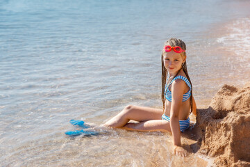 The girl in a blue swimsuit sits on the sand by the sea. She wears pink swimming goggles, and her...