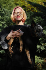 A young attractive woman with pink hair and glasses hugs with her Blue Heeler in spring park. Australian cattle dog on a walk with female owner. Family dog outdoor lifestyle concept.