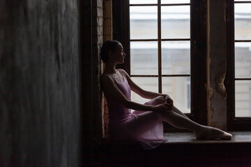 Ballerina in a pink dress and pointe shoes sits and looks out the window.