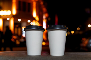 Close-up of two white eco-friendly recycled paper cups with coffee or tea standing on a bench against the background of a blurred night city with lights. Take the coffee with you.