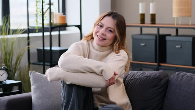 Portrait of a smiling and carefree Caucasian woman, looking at camera, sitting on sofa at home. Satisfied and cheerful lady spending leisure time.
