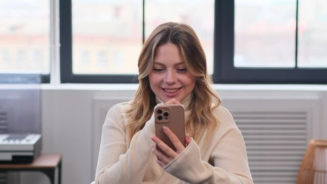 Friendly Caucasian young woman using phone and laughing while relaxing on sofa at home living room. Surfing internet and resting online.