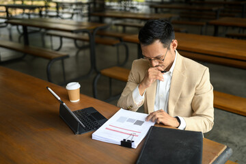 Focused asian male manager checking corporate financial documents at a cafeteria