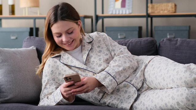 Happy Caucasian woman, lying on sofa at home living room, messaging in online social media using phone. Remote communication, internet leisure concept.
