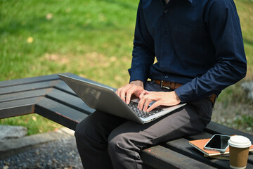 Smiling young businessman sitting on bench and using a laptop at public park