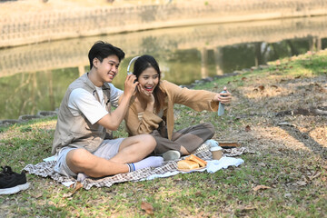 Cheerful young couple sitting on on blanket in park and taking selfie with smartphone