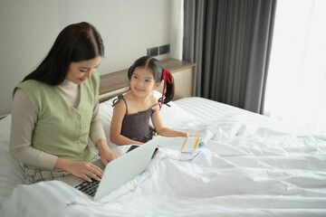 Beautiful mother working on laptop with daughter sitting on comfortable bed