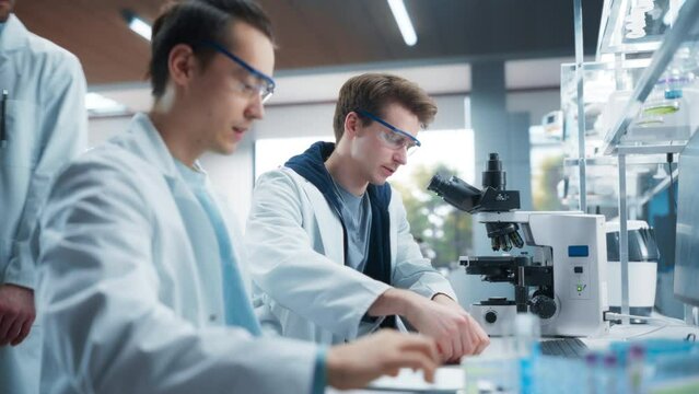 Two Young Medical Researchers Sitting Behind a Desk, Using Microscope for Medical Pharmaceutical Research and Treatment Development. Students Working on a Practical Assignment in a Modern Laboratory