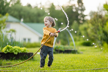 Cute little boy watering flower beds in the garden at summer day. Child using garden hose to water...