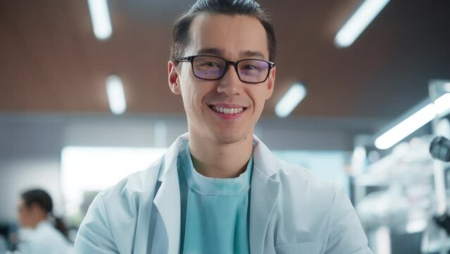 Portrait of an Asian Scientist Posing for Camera, Smiling. Young Male Biologist in a White Coat Working in a Modern Laboratory on a Laptop, Researching Medical Drug Products and Genome Editing
