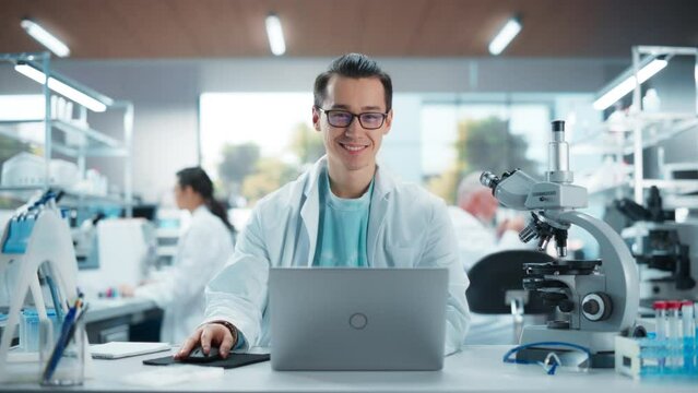 Genetic Research Scientist Working on a Laptop Computer in an Applied Science Laboratory. Portrait of a Handsome Young Multiethnic Lab Engineer in White Coat Looking at Camera and Smiling