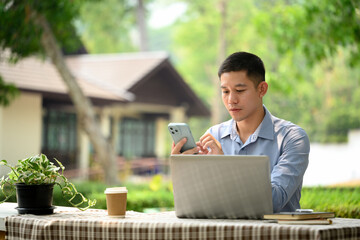Handsome male entrepreneur sitting outdoor with laptop and using mobile phone