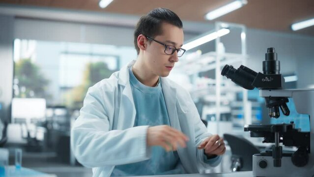 Cinematic Portrait of a Young Graduate Student Working in a Laboratory, Using Advanced Microscope, Writing Down Research Data on a Laptop. Multiethnic Biotechnology Specialist Using Modern Tools