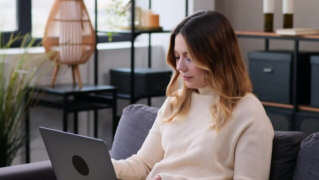 Serious Caucasian woman engaged in work or e-learning with laptop at home living room, sitting on sofa. Online education or entertainment, relaxation on free time.